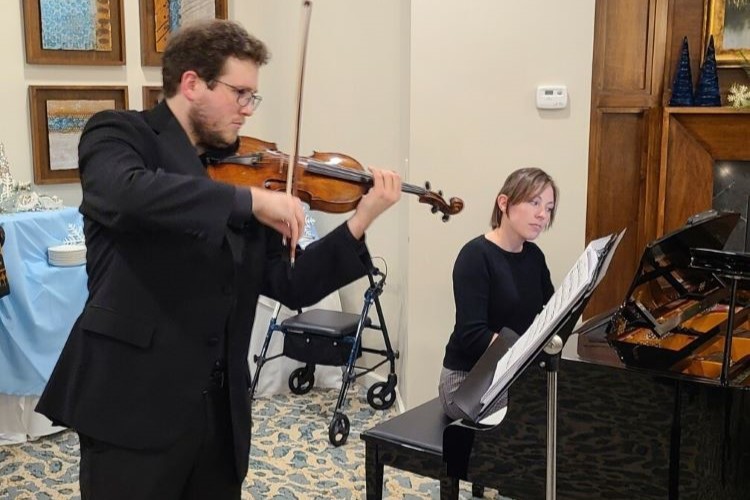 Beth plays a piano, a student plays the violin standing next to her