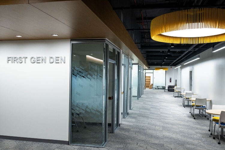 A sign says "First Gen Den" on the corner of a wall with collaborative space and couches to the side.